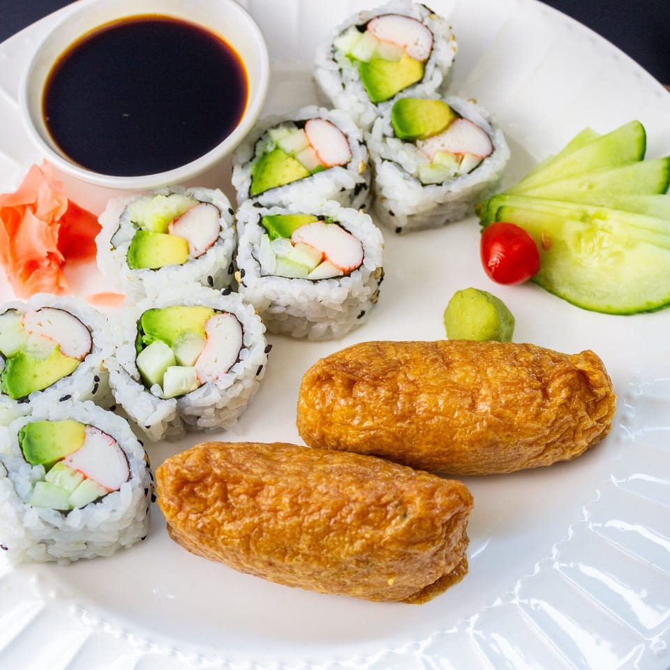 MAJÉ Sushi platter with California rolls, inari, soy sauce, pickled ginger, wasabi, and cucumber slices.